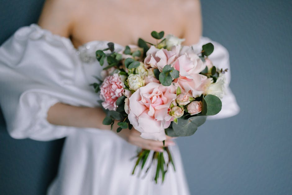 A person dressed in an elegant white off-shoulder gown holding a floral bouquet featuring large, soft pink garden roses, creamy white lisianthus, and green eucalyptus leaves. The bouquet is arranged i