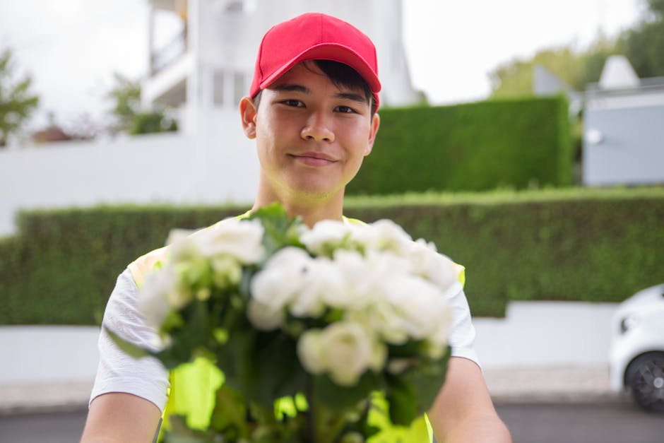 A young male flower delivery courier with tan skin, wearing a red cap and a high-visibility yellow vest, is smiling as he extends a bouquet of fresh white roses and lilies towards the camera. The bouq