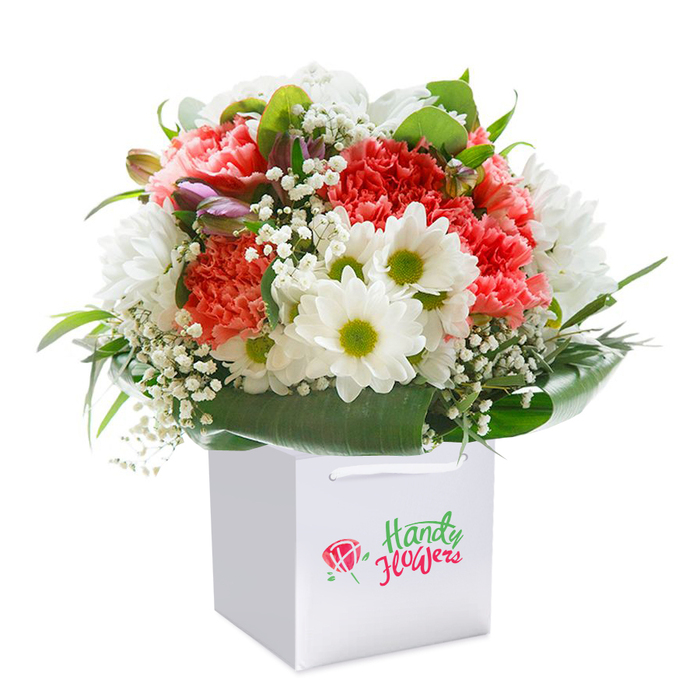 Captured against a seamless white background, a lush floral arrangement rises from a crisp, square white gift bag-an image evocative of the thoughtful craftsmanship found in Earlsfield's best local florists. The bouquet features a harmonious medley: abundant coral and soft peach carnations with intricately ruffled petals lend a luxurious texture and warmth, while snowy white daisies, distinguished by their bright lime-yellow centers, infuse the arrangement with a sparkling cheer akin to a riverside walk near the Wandle. Gossamer sprays of pure white baby's breath thread between the blooms, softening the composition and adding a subtle wildflower delicacy. Here and there, tightly closed purple-tinged buds nestle among the blossoms, suggesting quiet anticipation and a touch of seasonal intrigue, much like the first signs of spring in the Earlsfield neighborhoods. Shielding the floral centerpiece, broad, lacquered green leaves encircle the base, their deep colour and luster enhancing the bouquet's shape and imbuing it with a sense of fullness and professionalism. The bag's sturdy white ribbon handles arch gracefully overhead, designed for ease during local delivery or personal gifting. The overall presentation is immaculate and uplifting-crafted for joyful celebrations, thoughtful milestones, or a simple gesture to brighten someone's day in the heart of South West London. Each element suggests freshness and dedication, capturing the very essence of Earlsfield's flourishing floral scene.