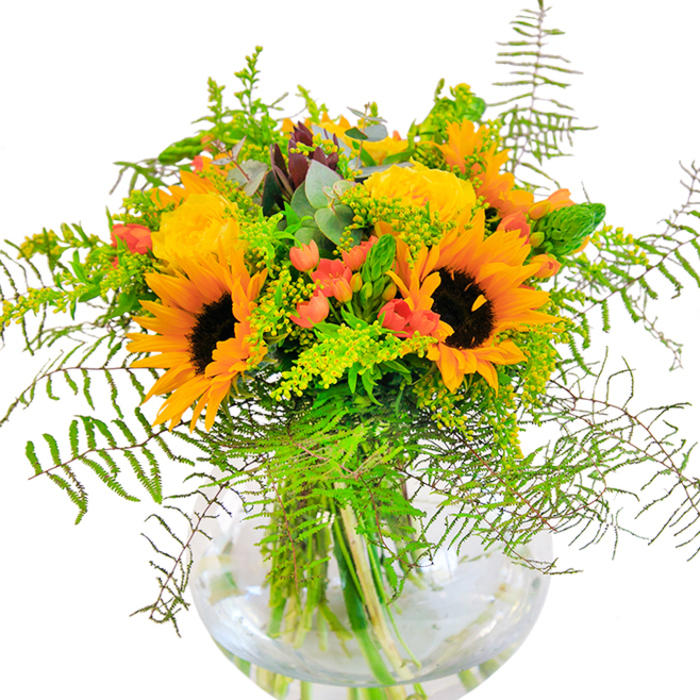 The essence of sunny Earlsfield afternoons is captured in this lush bouquet, where glossy yellow sunflowers and buttery garden roses gather in an artful, round glass vase. Each sunflower stands proud, its golden petals arching outward like rays from the heart of the arrangement; the rich, rust-brown centres invite warmth and familiarity, recalling the friendly faces seen along Earlsfield's streets. Nestled beside them, roses unfurl in perfectly formed spirals, their soft, velvety petals building layer upon layer of cheerful, golden hue. Interwoven throughout are gleaming clusters of scarlet Hypericum berries-jewels of colour that spark memories of local summer fêtes and family celebrations. Wisps of fern fronds, delicate and feathery, cascade gracefully, imparting a sense of gentle movement and untamed natural beauty. The bouquet's greenery is diverse-featuring airy light green solidago and hints of mellow eucalyptus, which offer fresh, herbal notes to the overall fragrance and evoke the wildness of the nearby Wandle Trail. The crystal-clear vase cradles the lush stems in cool water, a testament to freshness and the artisan's touch. As morning sunlight filters over the bouquet, shadows dance softly around each bloom, conjuring a sense of optimism and simple joy. Created for those cherished moments-an anniversary, a birthday, or a spontaneous act of kindness-this arrangement from Earlsfield Florist brings nature's exuberance and the neighbourhood's heartfelt energy to life.