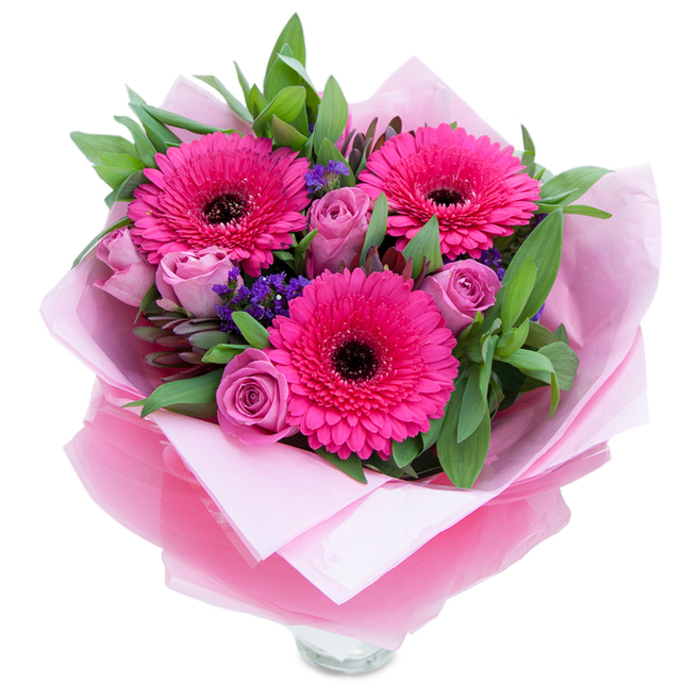 I see this arrangement as a playful, graceful study in pinks, captured simply against white so each bloom's character comes forward. Three fuchsia Gerbera daisies open like cheerful faces at the bouquet's core, their bright petals layered around a pronounced dark centre that gives the composition energy and contrast. Close beside them, roses in a dusty lavender-pink sit with petals still partly closed; their tight, velvety spirals promise a developing fragrance and a softness that balances the gerberas' boldness. Interspersed are sprays of tiny purple filler - statice or similar - whose feathery clusters add a whisper of texture and a cool counterpoint to the warm pinks. A generous backdrop of deep green foliage, the kind we select for structure and longevity (salal or eucalyptus-style leaves), creates a lush, garden-like base and a sense of botanical richness. The bouquet's wrapping has been chosen to echo the palette: an inner layer of pale pink tissue for a tender, matte base and an outer sheet of translucent fuchsia-pink cellophane that shimmers slightly and folds neatly around the stems. The clear glass vase peeks beneath, its rim visible and suggesting the flowers are arranged for immediate display, whether on a dining table or a bedside in an Earlsfield flat. Under soft daylight the textures and hues read as celebratory and comforting; the bouquet feels like a deliberate, hand-crafted gesture - perfect for marking a birthday, anniversary, or simply saying 'you're thought of' to someone in Wandsworth borough.