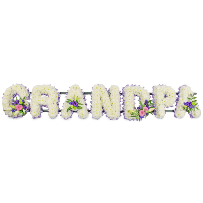 A detailed funeral flower tribute in the shape of the word "GRANDPA" stretches horizontally across the frame, set against a simple white background that highlights every nuance of colour and texture. Seven capital letters, each upright and substantial, are completely filled with compact white chrysanthemums or similar classic sympathy blooms, their many layered petals creating a dense, velvety surface that looks soft to the touch. A narrow edging of lavender and soft purple flowers traces around each letter, lending a gentle contrast and helping the shapes stand out clearly while still keeping the palette calm and dignified. Within selected letters, thoughtfully placed accent sprays introduce subtle movement and richer tones: the 'G' carries a small bouquet of white lily-style flowers accompanied by vivid pink and fuchsia heads and a touch of greenery; the first 'A' and the last 'A' bear smaller groupings of purple blossoms nestled into the white; and the 'D' features a fuller spray echoing the 'G', with pink blooms, a graceful white lily, and glossy green leaves. A slim, dark green bar or frame runs discreetly along the rear, giving the impression that the letters are mounted on a stand, as you might see at a memorial service in Earlsfield or at a nearby chapel in Wandsworth. The even lighting and clean setting draw attention to the craftsmanship of the tribute-the way each flower head is placed to form a perfectly legible, heartfelt message. It feels like a deeply personal way to honour a beloved grandfather's memory, with a balance of purity, colour, and quiet respect.